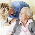 Care worker making a home visit. Female carer is hoovering the living room to help an elderly woman. The woman is sitting on a sofa relaxing