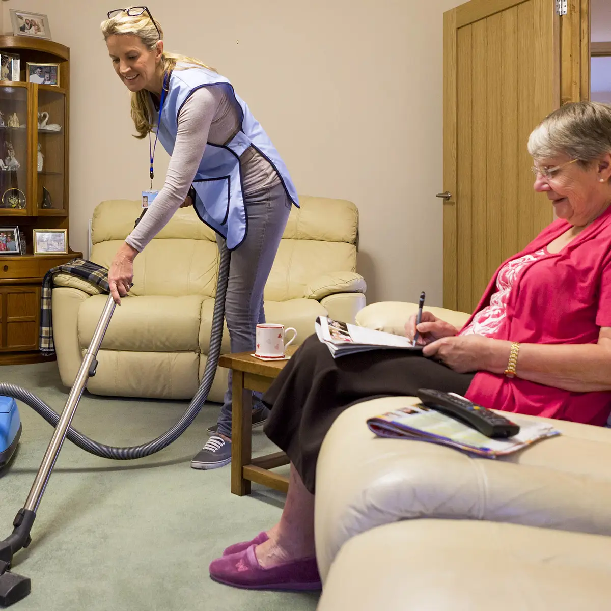 Care worker making a home visit. Female caregiver is hoovering the living room to help an elderly woman. The woman is sitting on a sofa relaxing