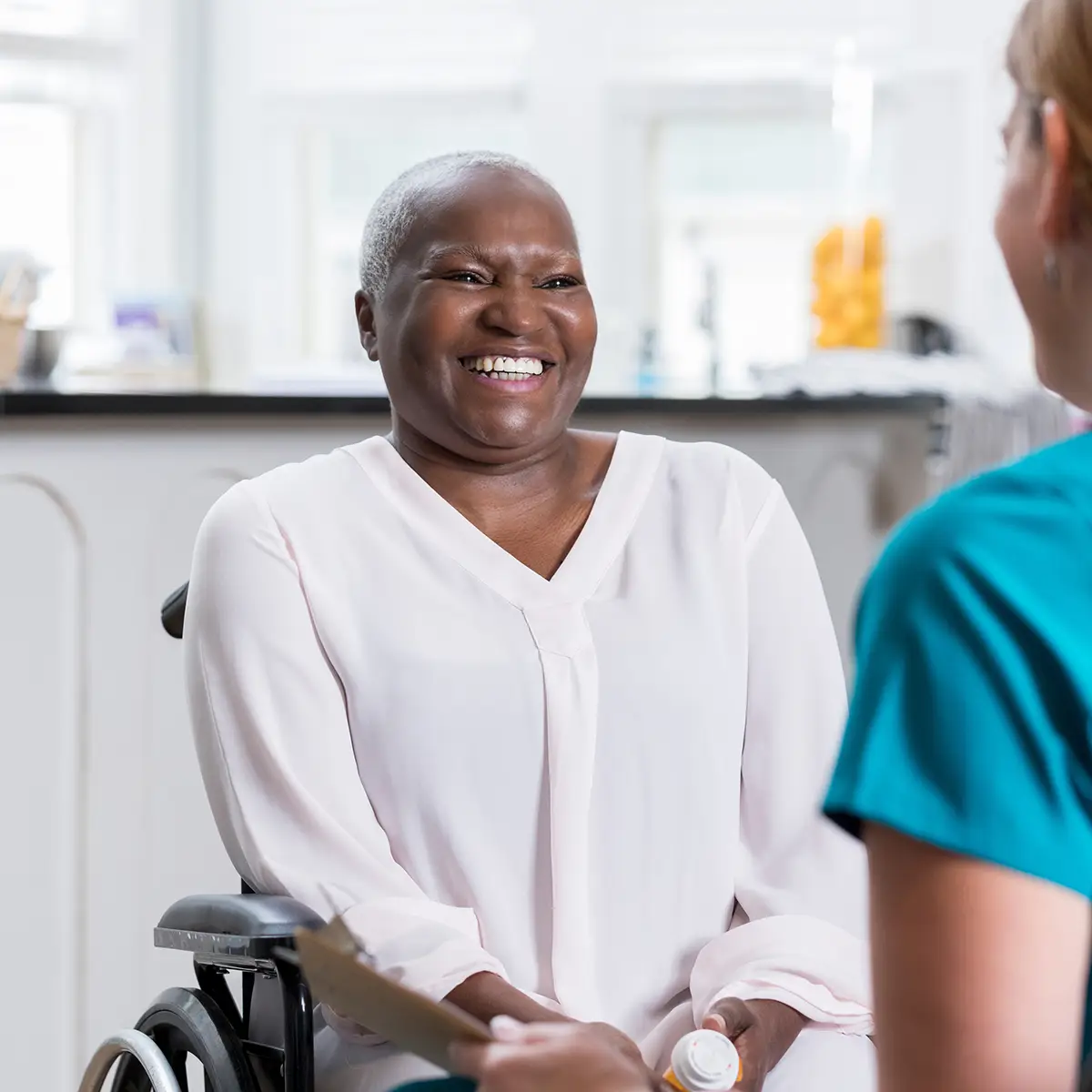 Beautiful African American senior woman smiles and laughs while talking with her unrecognizable home healthcare nurse. The patient is sitting in a wheelchair.